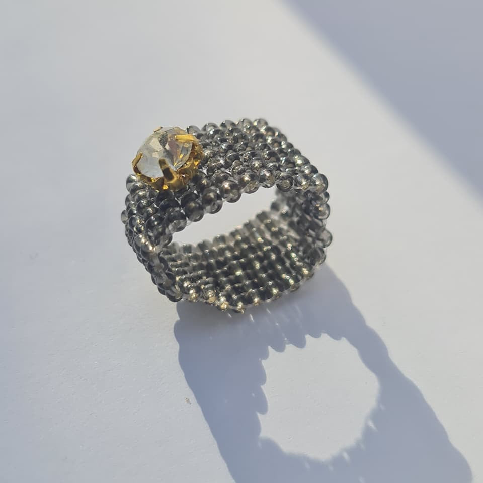 A beaded ring with a textured dark gray beaded band and a clear faceted crystal accented with a gold setting, resting on a white surface with a soft shadow.