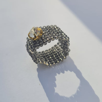 A beaded ring with a textured dark gray beaded band and a clear faceted crystal accented with a gold setting, resting on a white surface with a soft shadow.