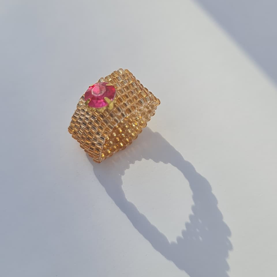 A gold ring with a textured honeycomb pattern and a small pink flower accent on top, photographed on a white surface with a sharp shadow.