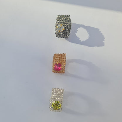 Three small square jewelry pieces arranged vertically on a white surface, each with a textured metal border and a central gemstone in different colors: silver with a clear stone, gold with a pink stone, and white with a yellow-green stone.