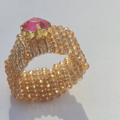 A handmade beaded ring featuring a textured, multi-row design of small gold and clear beads, topped with a faceted pink gemstone in a gold setting, photographed on a white surface with a soft shadow.