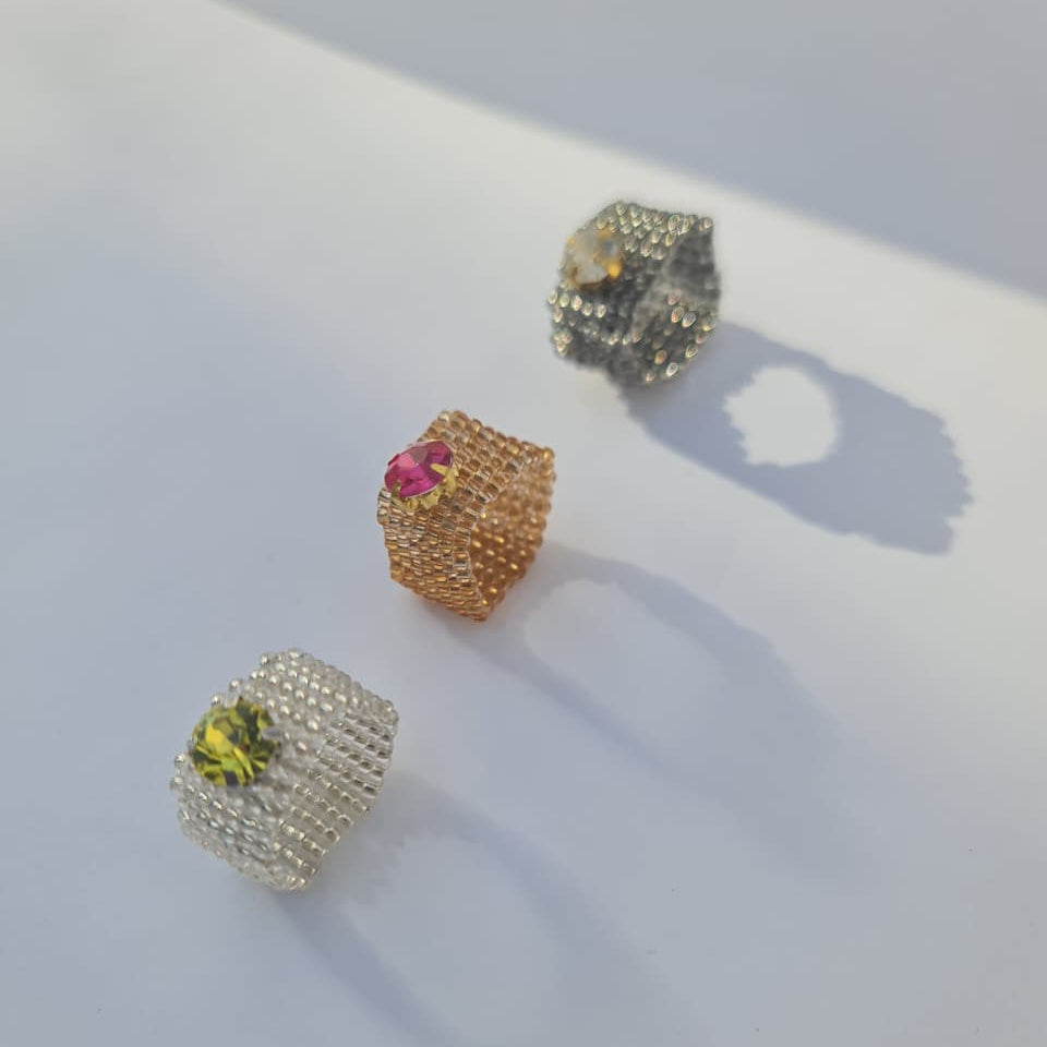Three textured cube-shaped rings with small pavé settings and central gemstones, displayed on a white surface. One ring has a yellow stone, one has a pink stone, and one has a clear stone set in a silver-toned metal.