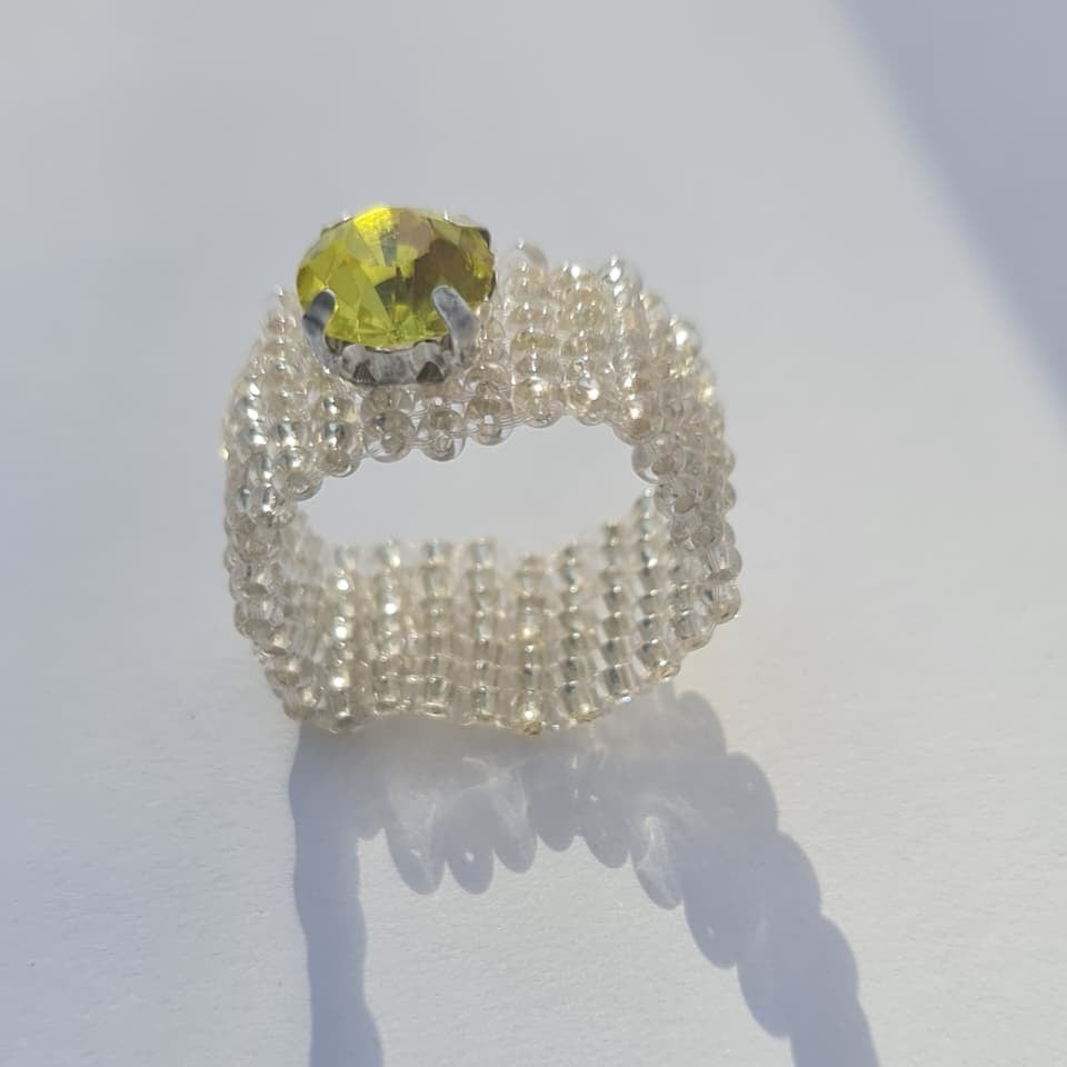 A handmade beaded ring featuring a textured, multi-strand silver band and a round yellow-green gemstone, displayed on a white surface with a soft shadow.