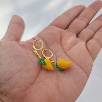 A pair of small gold hoop earrings with yellow fruit charms and green leaf details, resting on an open hand against a bright white background.