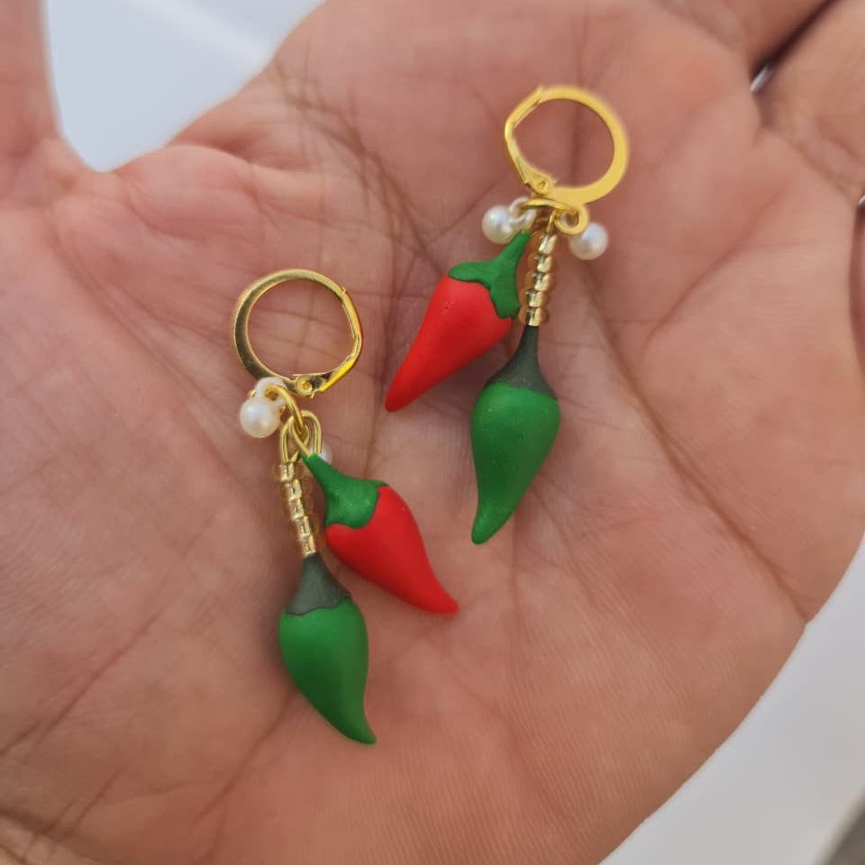 A pair of chili pepper-shaped earrings with gold-tone rings, green stems, and small white pearl-like beads, resting on an open hand.