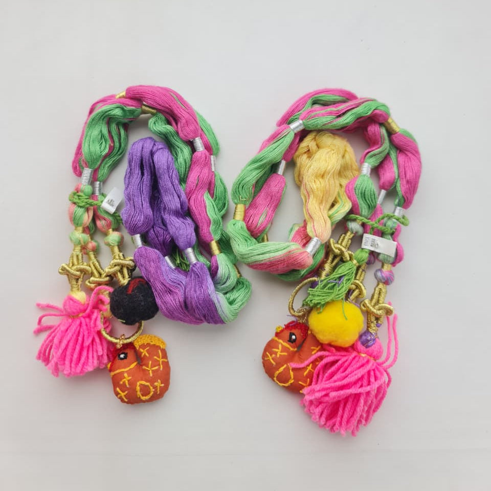 Two colorful braided hair bundles with tassels and decorative charms, displayed on a plain white surface. The hair is woven with pink, green, purple, and yellow threads, and features small embroidered character charms.