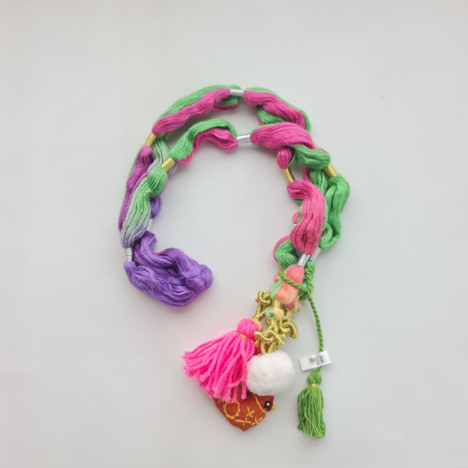 A handmade braided hair accessory made of pink and green thread, featuring a purple tassel, a pink tassel with a small gold heart charm, a white pom-pom, and a green tassel, laid on a white surface.