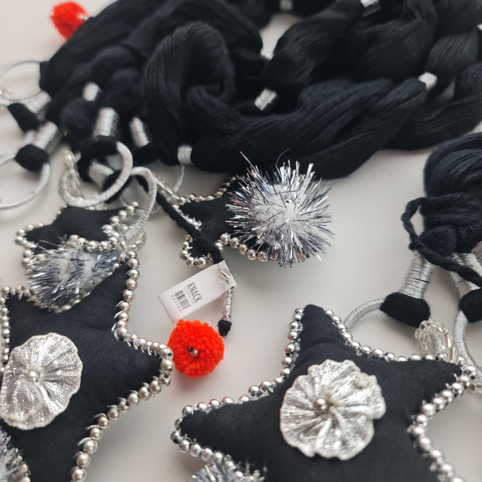 Black fabric star-shaped ornaments with silver beaded edges and silver flower appliqués, shown with black tassels and small red and silver pom-poms on a light background.