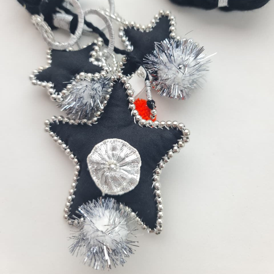 A close-up of a handmade black fabric star pendant necklace with silver beaded edges, silver tinsel pom-poms, and a small red accent, shown on a white background with black yarn in the upper right.
