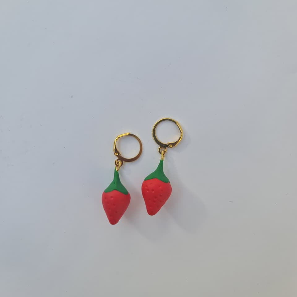 A pair of small gold hoop earrings with dangling strawberry charms, featuring textured red bodies and green tops, set against a plain light gray background.