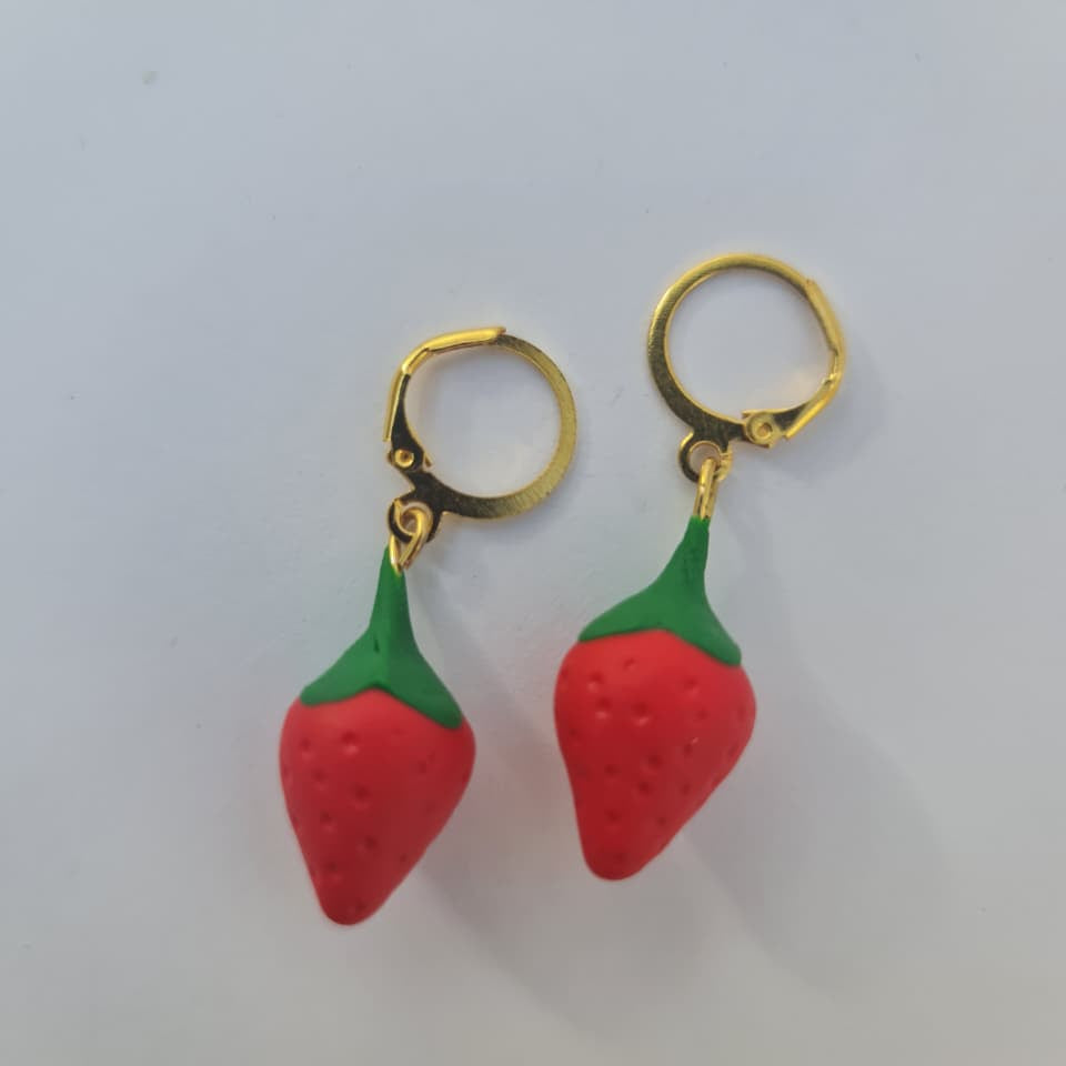 A pair of gold hoop earrings with dangling red strawberry charms that have green tops, shown on a plain light gray background.