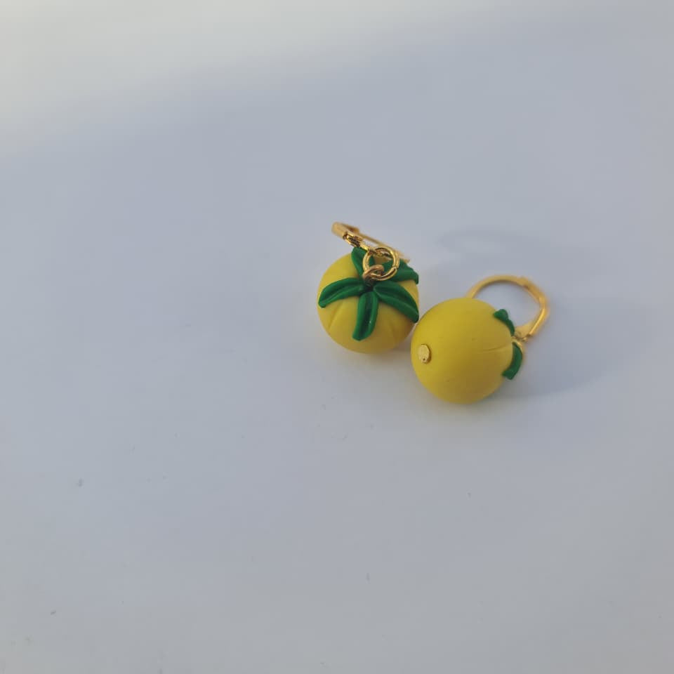 A pair of small yellow lemon-shaped earrings with green leaf accents and gold-tone rings, photographed on a plain white surface.
