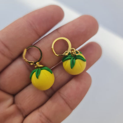 A pair of small yellow fruit-shaped hoop earrings with green leaf details and gold-tone hoops, held in a person's hand against a light background.