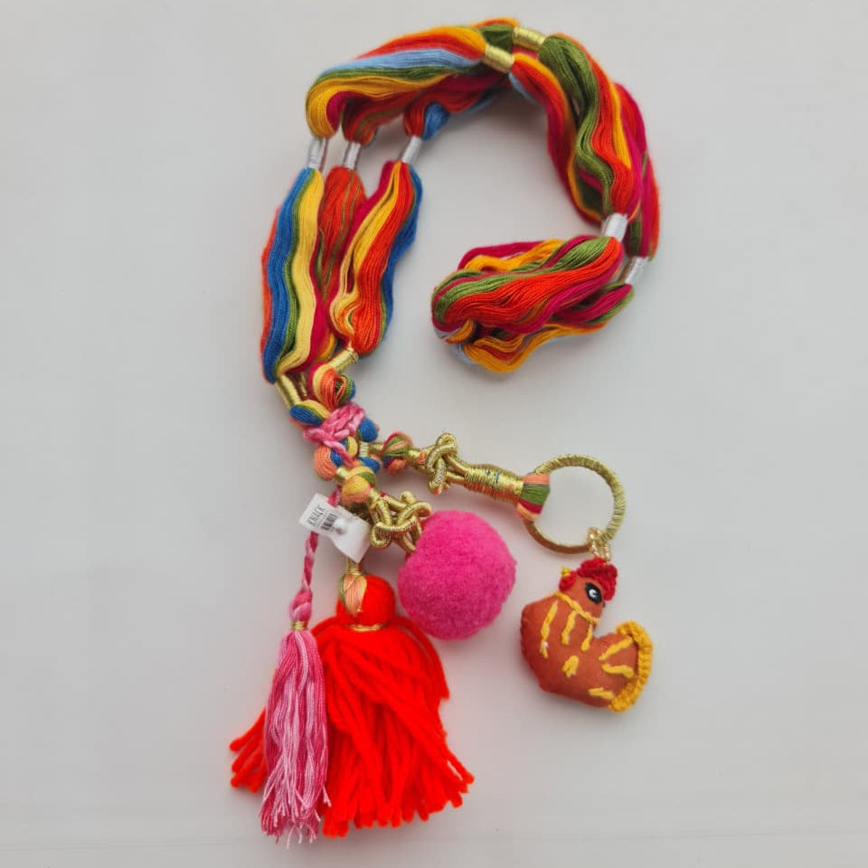 A multicolor woven cord bracelet or lanyard with tassels, a pink pom-pom, and a small embroidered rooster charm, displayed on a plain light background.