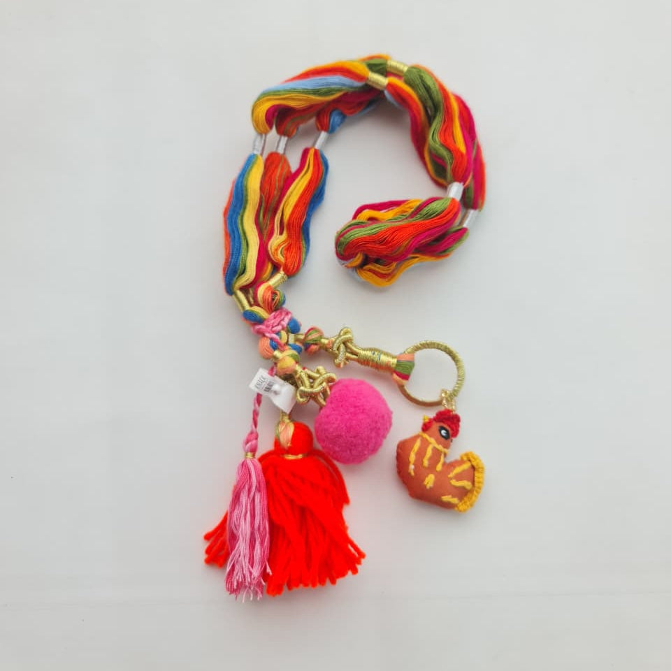 A multicolor braided cord keychain with a gold-tone clasp, featuring a red tassel, a pink pom-pom, and a small orange and yellow bird charm, displayed on a plain white background.