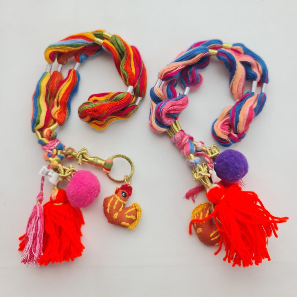 Two colorful braided cord keychains with gold-tone hardware, tassels, pom-poms, and small decorative charms, displayed on a plain white background.