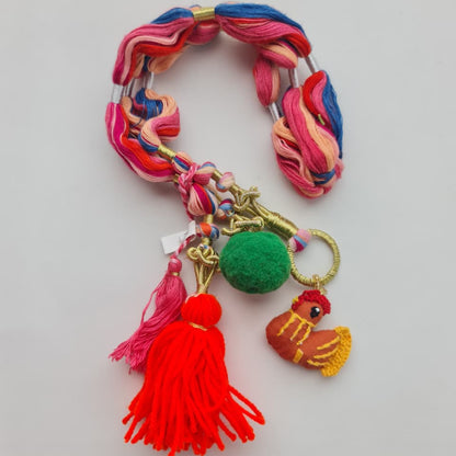 A handmade multicolor braided cord bracelet or keychain charm featuring pink, blue, and orange yarn, gold-tone metal rings and clasps, a green pom-pom, and a small embroidered rooster charm, displayed on a light gray background.