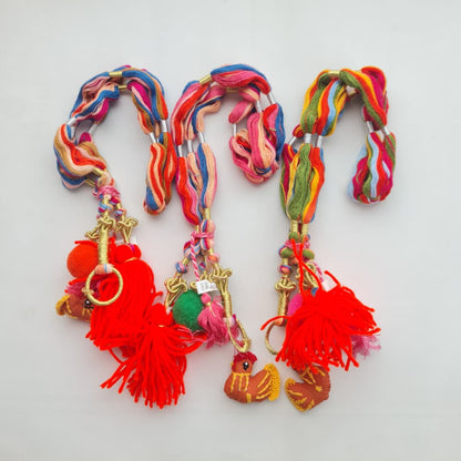 Three colorful braided hair ties with tassels and small decorative charms, displayed on a white sheet of paper.