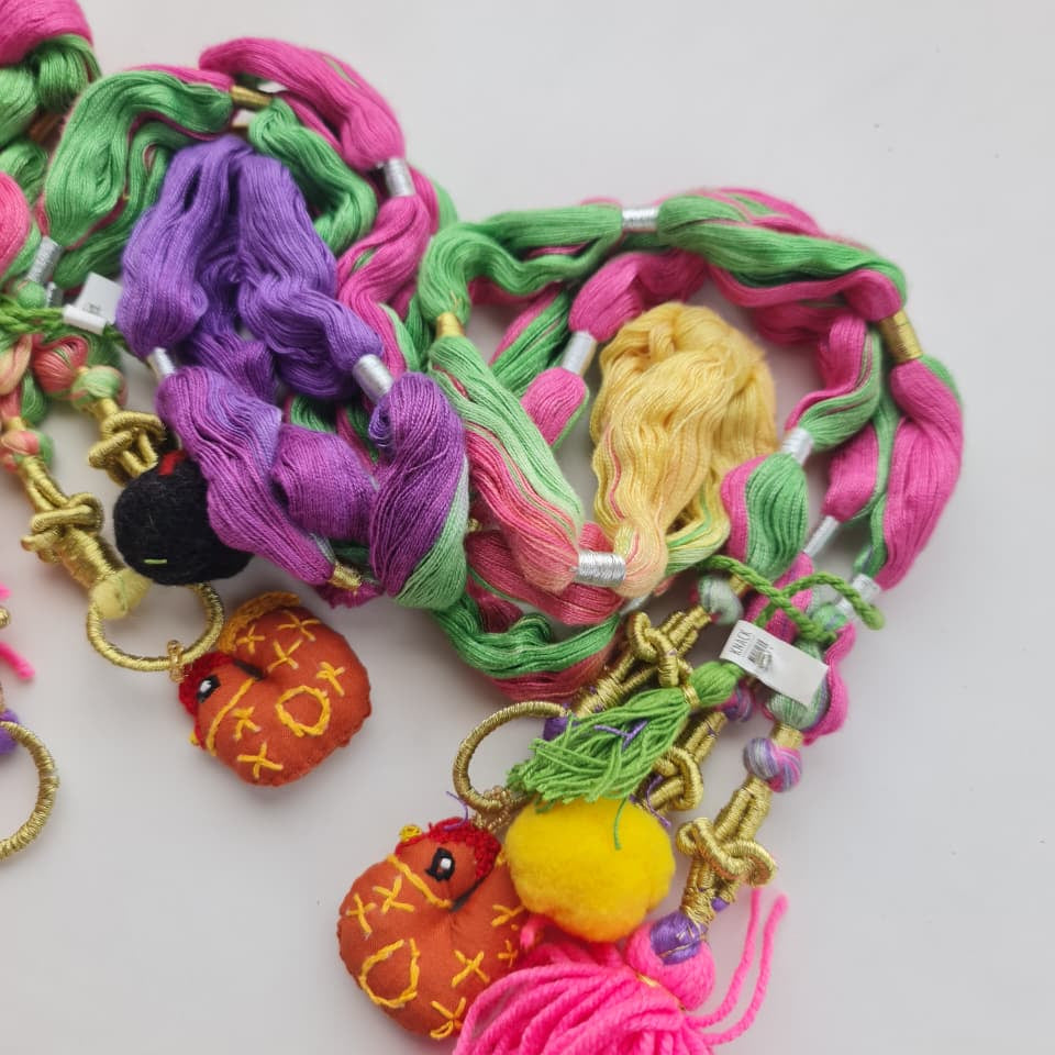 A close-up of a colorful braided hair accessory with pink, green, purple, and yellow braid strands, gold-tone metal rings, small beads, and decorative tassels and fruit-shaped charms on a plain light background.