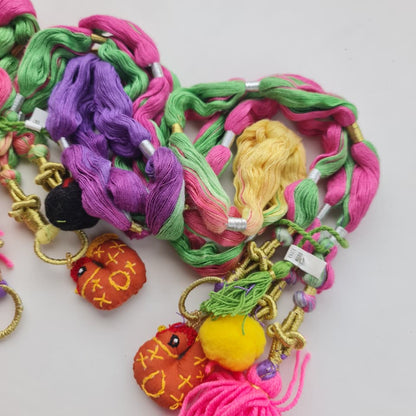 A close-up of a colorful braided hair accessory with pink, green, purple, and yellow braid strands, gold-tone metal rings, small beads, and decorative tassels and fruit-shaped charms on a plain light background.