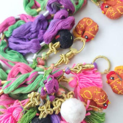 A close-up of a colorful handmade tassel charm or keychain featuring bright pink, purple, green, yellow, and orange threads, gold-tone metal rings and beads, and small embroidered bird embellishments on a white background.