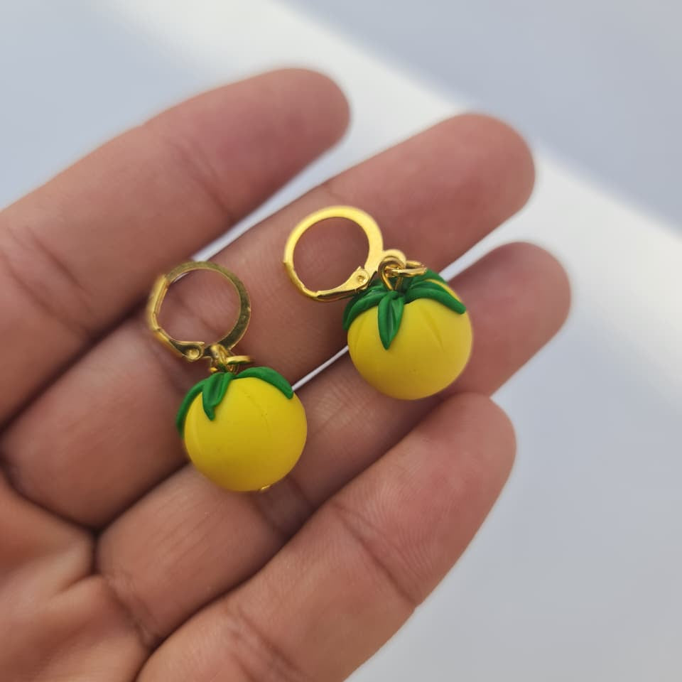 A pair of small yellow lemon-shaped hoop earrings with green leaf details and gold-toned hoops, resting on a person's palm against a light background.