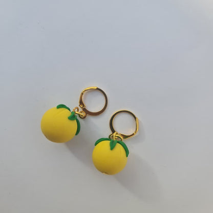 A pair of gold hoop earrings featuring small yellow lemon charms with green leaf accents, displayed on a plain light gray background.