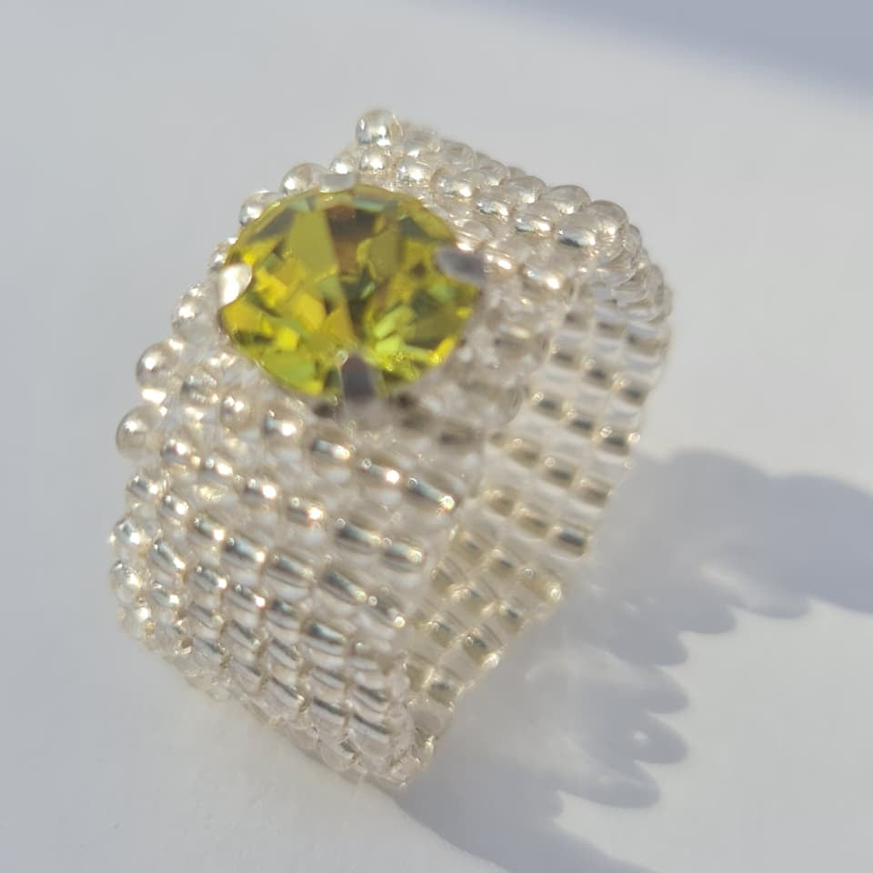 A handmade beaded ring featuring a large, faceted yellow-green gemstone in the center, surrounded by textured silver and white seed beads on a plain white background.