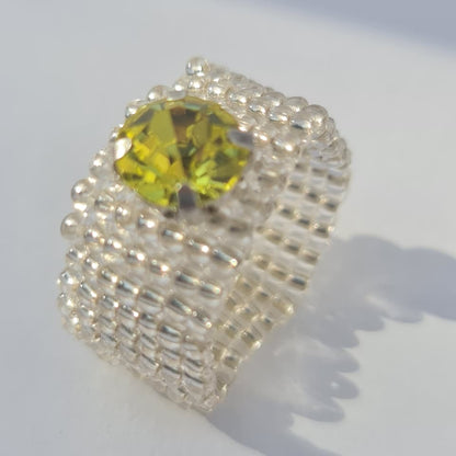 A handmade beaded ring featuring a large, faceted yellow-green gemstone in the center, surrounded by textured silver and white seed beads on a plain white background.