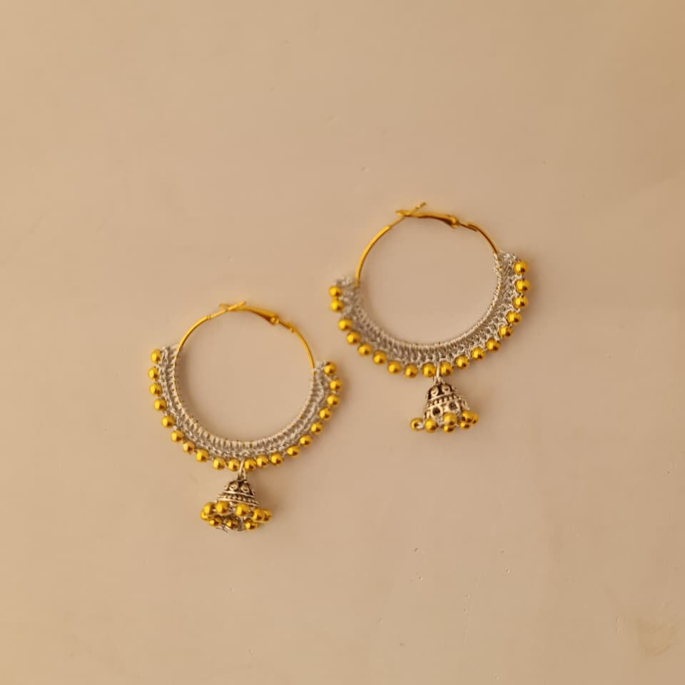 A pair of gold and silver hoop earrings with small dangling bell charms, displayed on a solid beige background.