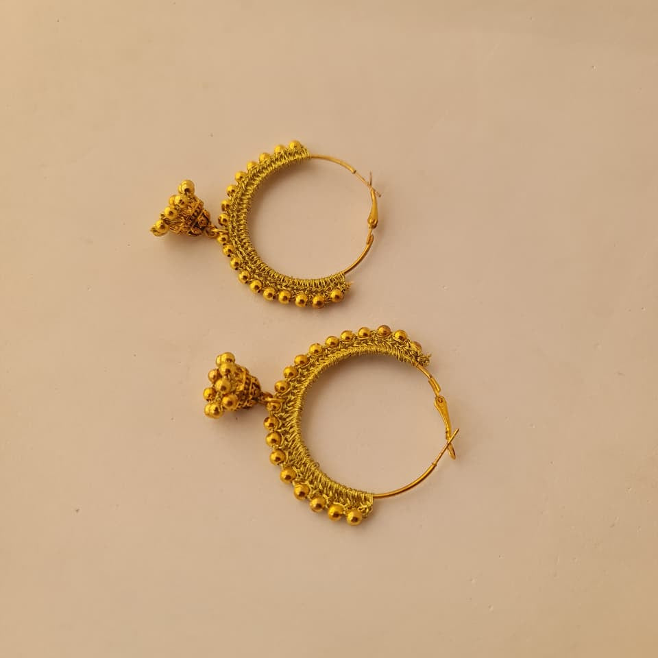 A pair of gold-toned hoop earrings with intricate textured detailing and small dangling elements, displayed on a plain beige background.