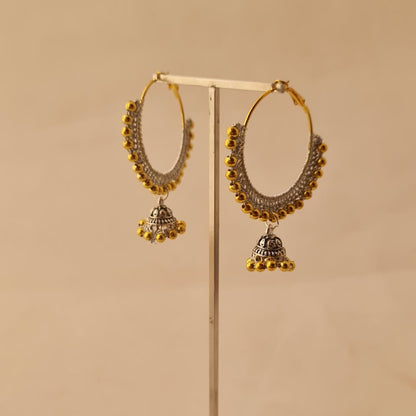 Gold and silver hoop earrings with dangling bell-shaped charms and beaded fringe, displayed on a metal stand against a warm beige background.