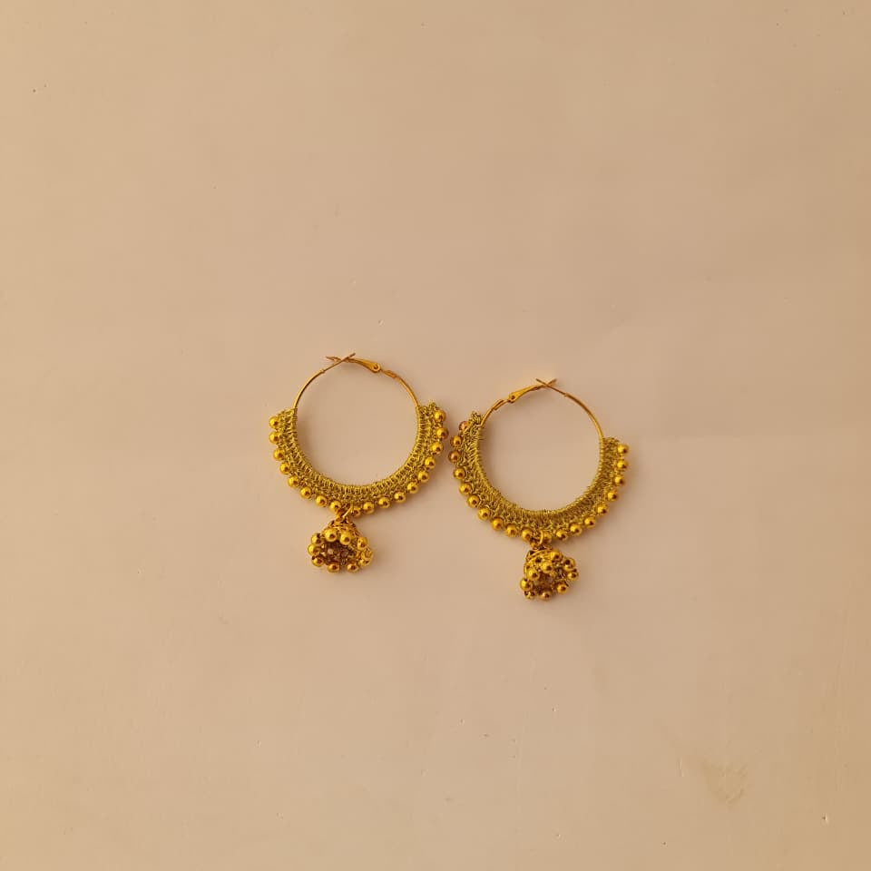 A pair of gold-toned hoop earrings with textured beaded edges and small dangling heart-shaped charms, displayed on a plain beige surface.