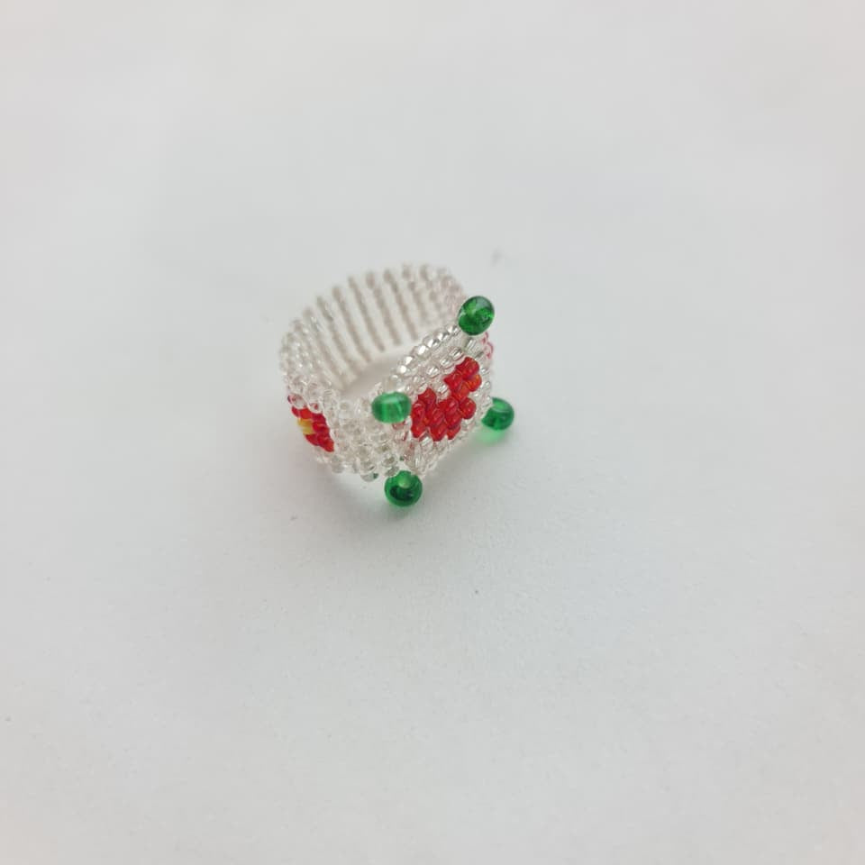 A handmade beaded ring featuring a white textured band decorated with red and green beads, photographed on a plain white background.