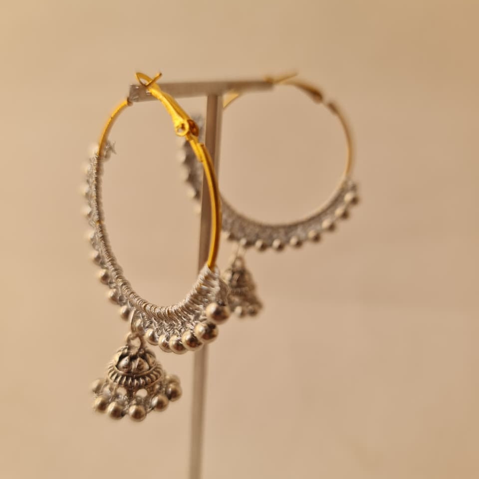 Silver-toned hoop earrings with dangling jhumka-style charms, displayed on a stand against a plain beige background.