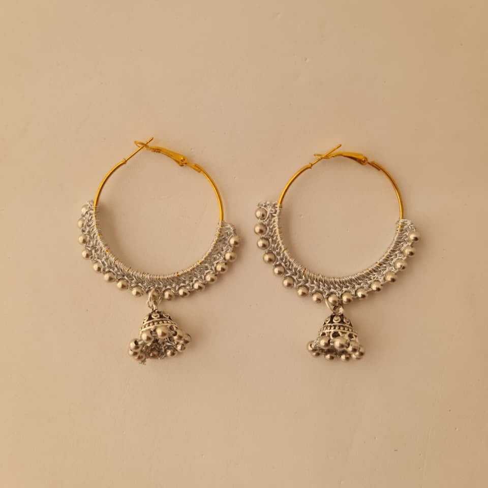 A pair of gold and silver-toned hoop earrings with intricate beadwork and dangling bell-shaped charms, displayed on a plain beige background.