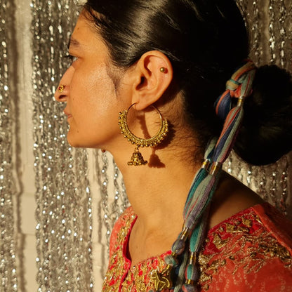 A woman in profile wearing large gold-toned hoop earrings with dangling bells, a red embroidered top, and a blue braided hair accessory, standing in front of a silver beaded curtain.